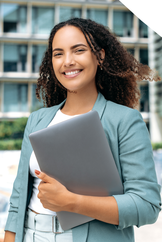 Smiling person with curly hair in a teal blazer holds a closed laptop outdoors, with a modern building in the background.