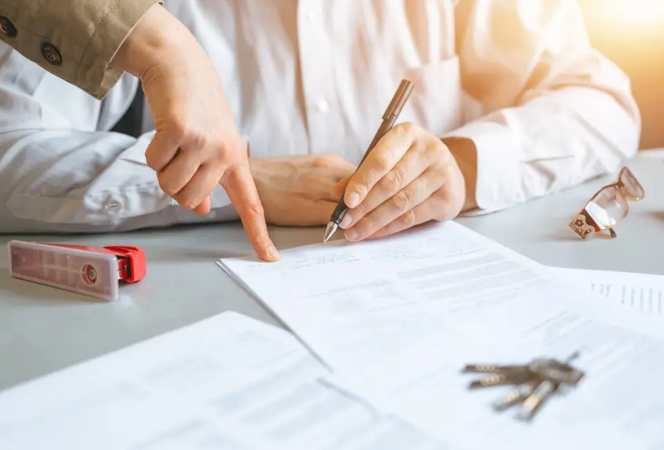 A person signs a document at a desk while another points at it. Nearby are a pair of glasses, a tape measure, and keys.