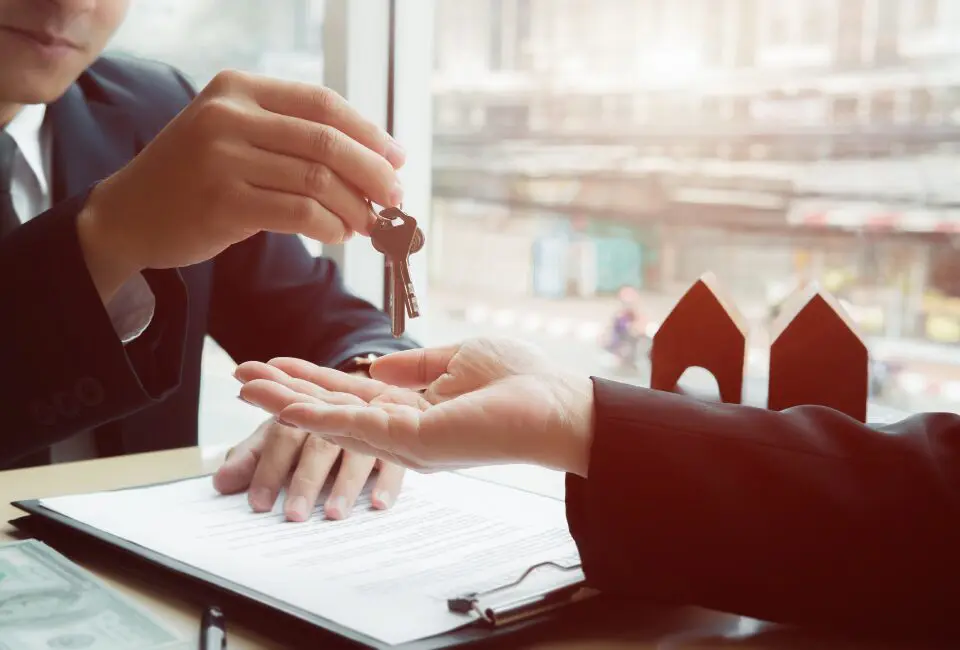 A person hands keys to another across a table with a contract and money, suggesting a property deal or rental agreement.